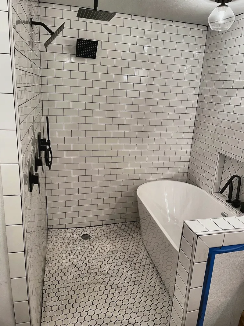 The master bathroom tub and shower area during tiling, with the freestanding tub set and painter's tape still on the curb